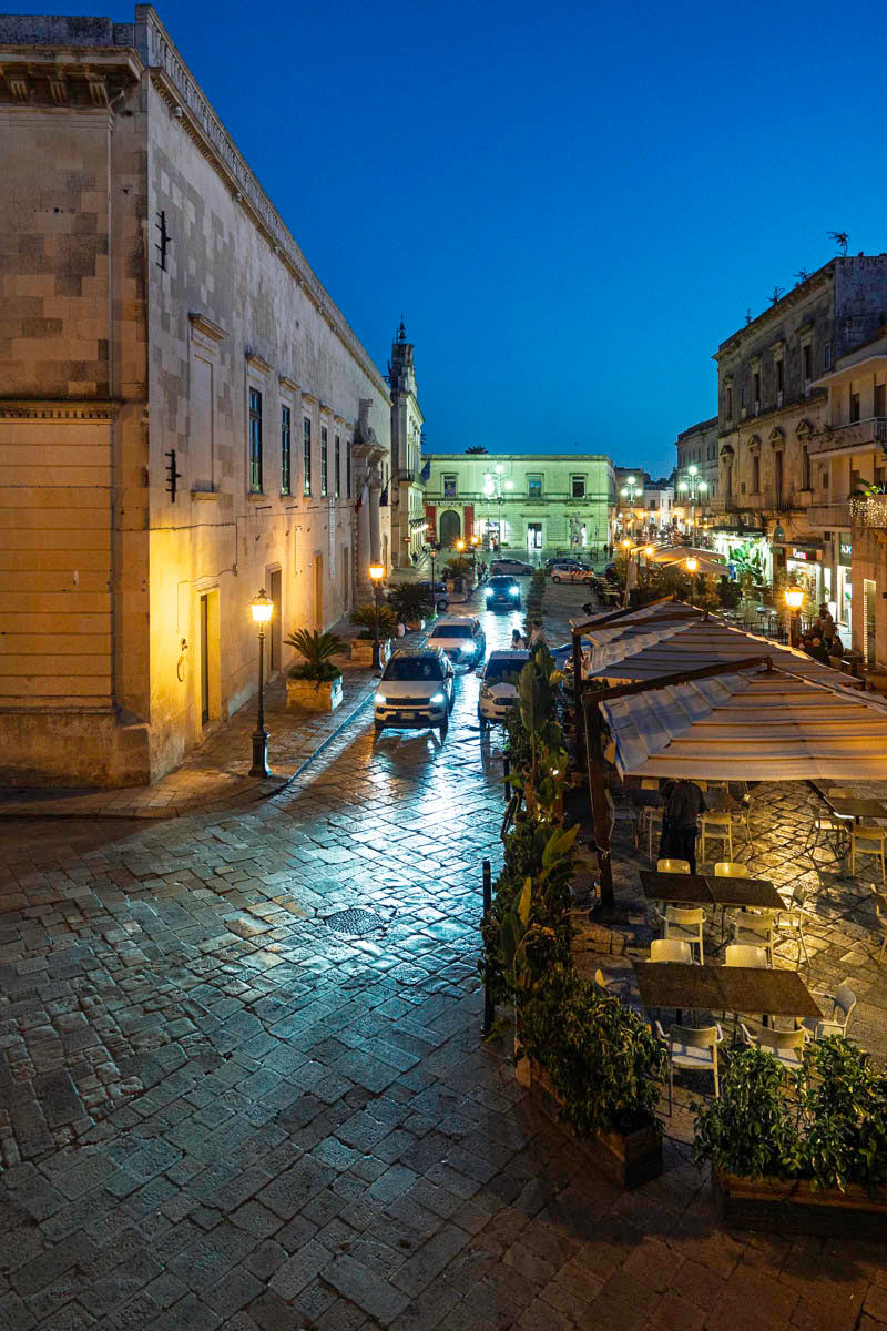 A Maglie street at blue hour, with café terraces and wet paving reflecting the street lights