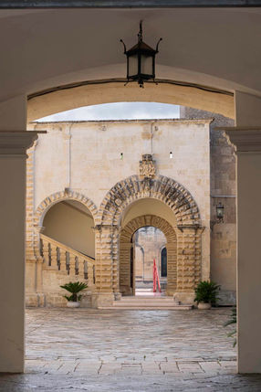 A view through an arch into a stone courtyard with a carved double arch and staircase in Maglie