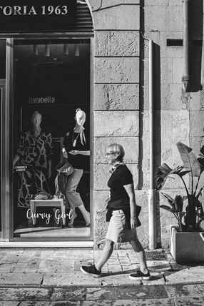 Black and white photograph of a woman walking past a shop window with two mannequins in Maglie