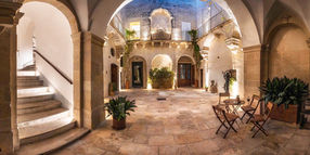 A wide view of the lamplit central courtyard of Palazzo Garzia in Maglie, with a stone staircase, arches and café tables