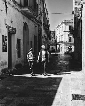 Black and white photograph of two boys walking through a band of sunlight in a narrow Maglie street