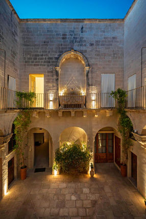 The central courtyard of Palazzo Garzia in Maglie lit at blue hour, with a carved niche, balconies and planted arches