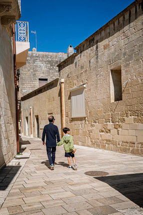 A man and a boy walking hand in hand along a sunlit limestone street in Maglie