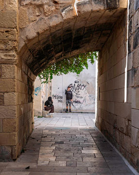 A stone archway in Maglie leading to a sunlit wall with graffiti and two figures