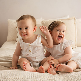 Dos bebés felices sentados en la cama, sonriendo. Infantil/Familia, contexto hogareño, luz natural.