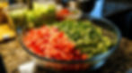 A glass bowl with diced tomatoes and guacamole on a granite countertop, surrounded by bowls of chopped veggies in a well-lit kitchen.