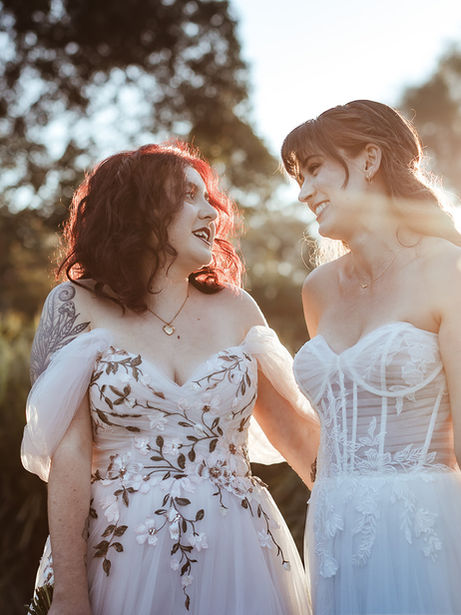 Two beautiful brides with a stunning sunset behind them, smiling at each other on their wedding day.