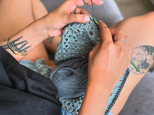 Close up image of author crocheting a piece in teal and grey. Her self designed and applied tattoos are visible on her arms and legs.