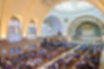 Large congregation seated in a colorful church with stained glass windows, arched ceiling, and ornate altar; serene and reverent atmosphere.