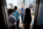 People at a door, exchanging a grocery bag with a man from the SVDP Society. Smiling faces, overcast day. Sparse trees and parked cars in the background.