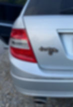 Wir sehen das linke Rücklicht eines silbernen Autos. Rechts daneben hängt ein magnetischer, 2,5 dimensionaler catcar Schriftzug. Die Trägerplatte des Schriftzuges ist schwarz und die Oberfläche in irisierenden Metallflakes mit einer glänzenden Harzoberfläche.