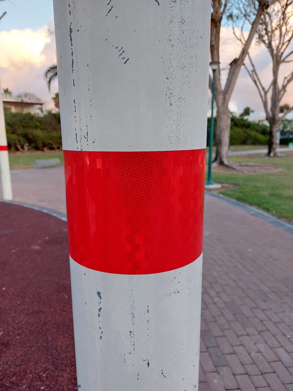 "עמוד לבן עם סרט מחזיר אור בצבע אדום, מותקן בגובה נגיש בסביבה ציבורית עם מדרכה ודשא ברקע. מחזיר האור משמש להגברת הנראות והבטיחות, במיוחד עבור אנשים עם לקות ראייה והולכי רגל."