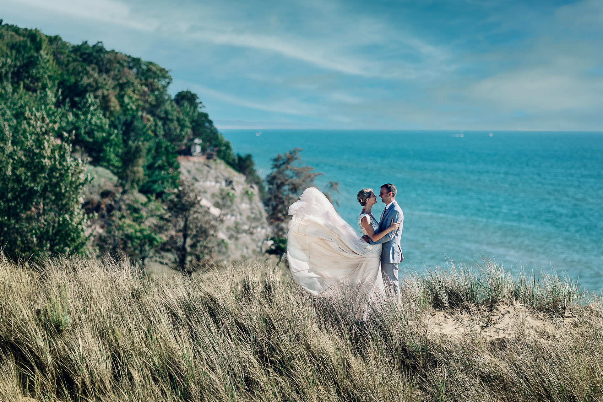 Couple on sand dune with woods and Lake Michigan in. the background Her dress is flowing perfectly in the wind.