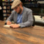 Autor branco, jovem, com barba e óculos, de perfil sentado em uma cadeira, autografando um livro em uma grande mesa de madeira. Atrás dele tem várias estantes de livros.