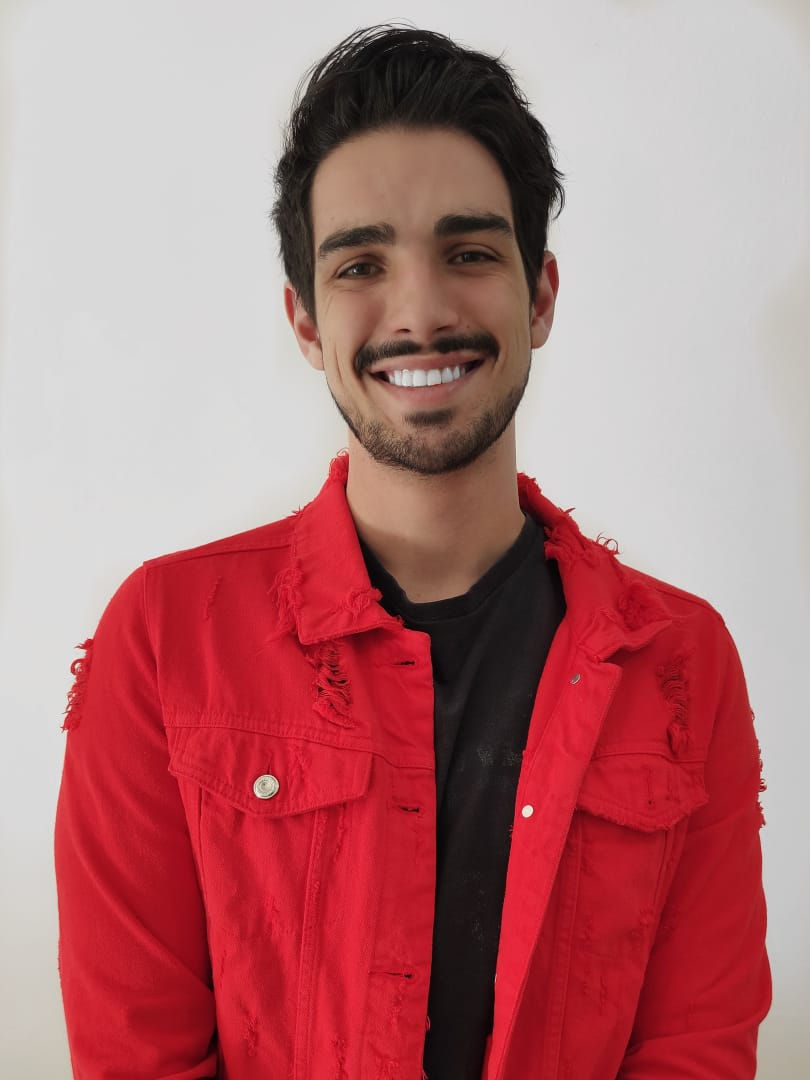 Homem branco, sorrindo, com uma camisa preta e jaqueta vermelha. Cabelos escuros e tem bigode e barba.