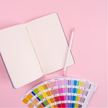 Fundo rosa claro, com um caderno aberto com páginas brancas um lápis em cima e uma paleta de cores em baixo.