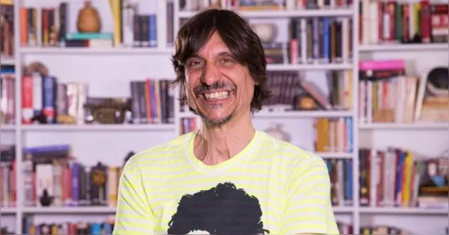 Peninha sorrindo em plano americano com estande e livros de fundo