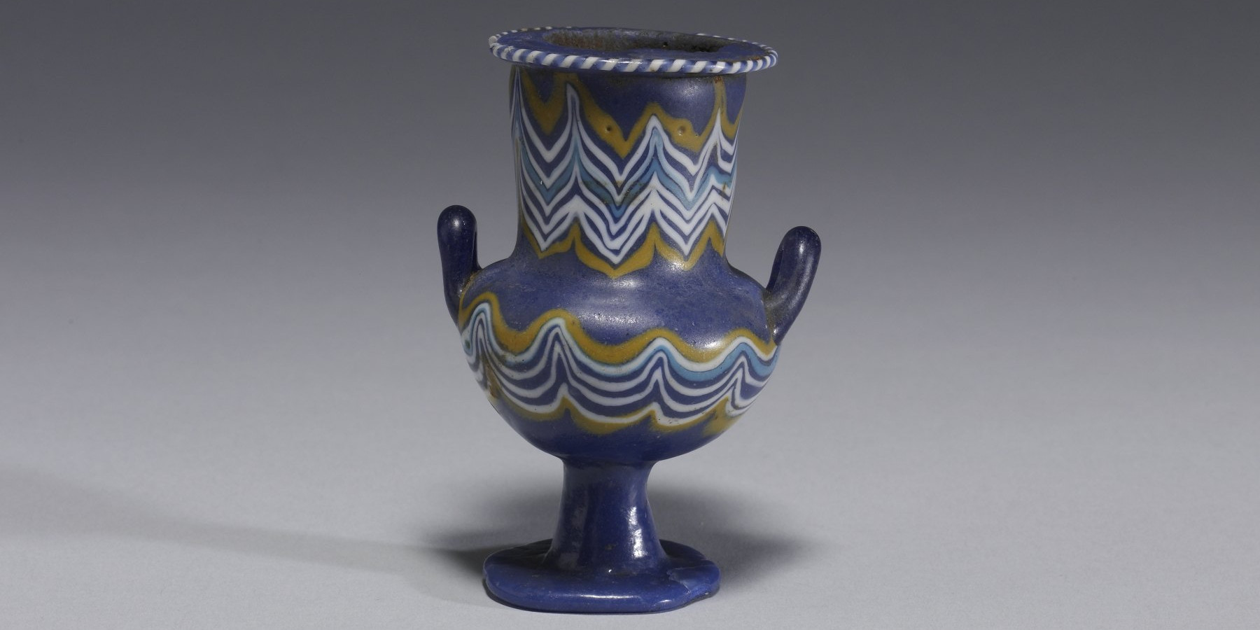 Small ancient Egyptian glass vessel, opaque blue with yellow and white wavy trails, two handles