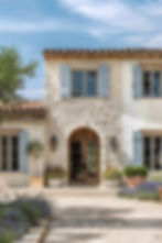 Mediterranean-style house with stone walls, blue shutters, arched wooden door, and potted plants.