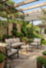 Modern patio with beige sofas, plants, and a pergola. Stone wall and greenery create a serene outdoor setting.