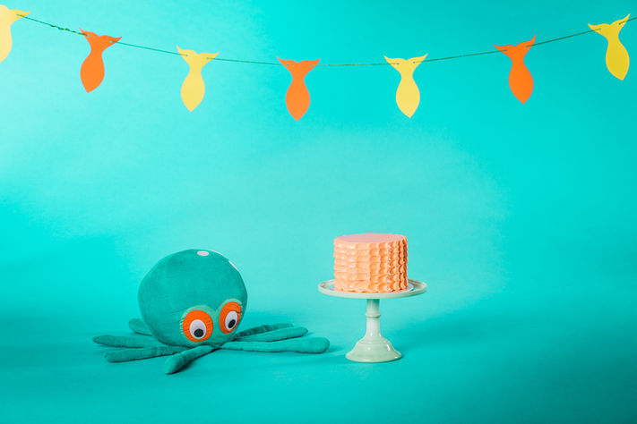 Studio shot of orange gourmet cake on a stand beside a plush toy octopus under fish pennant
