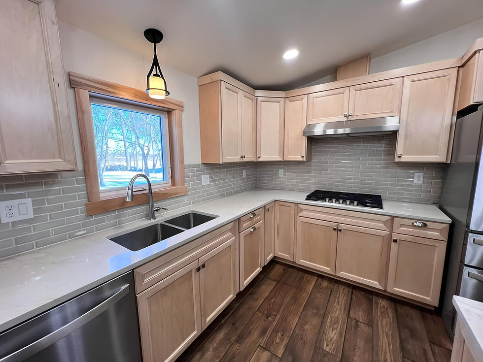 Remodeled kitchen featuring natural maple cabinets, white quartz countertops, a double-basin stainless steel sink, and a stainless steel gas cooktop with matching range hood. The backsplash is a light gray subway tile, and the flooring is a rich, wood-look vinyl plank. A large window framed in wood trim brings in natural light, with a pendant light hanging above the sink.