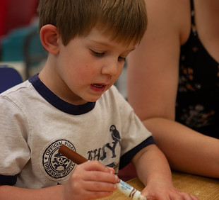 Young boy carefully uses a crayon to draw on yellow playdough at school.