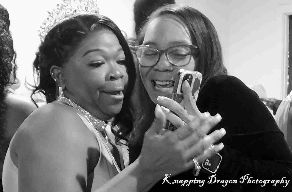 Two women share a joyful moment over a phone at an event, beautifully captured in black and white by Knapping Dragon Photography.