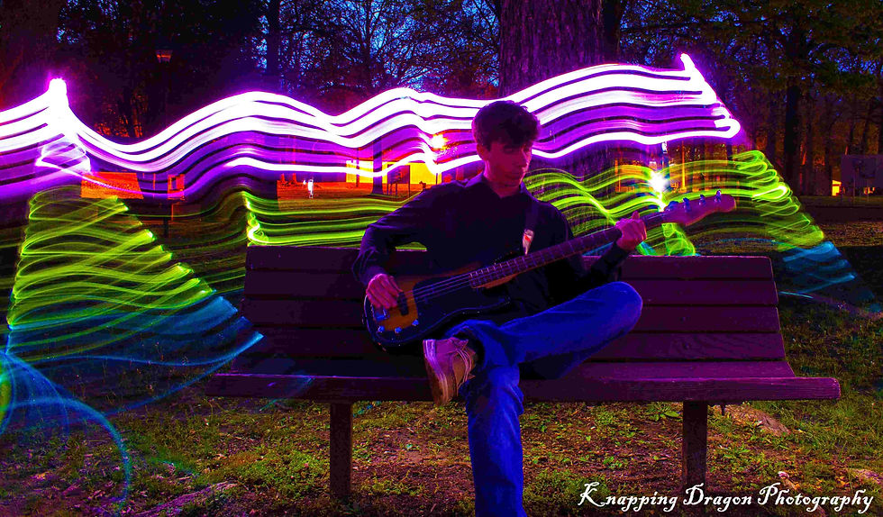 A guitarist sits on a park bench, enveloped in vibrant swirls of purple and green light trails, creating a mesmerizing composition in this light art photograph by Knapping Dragon Photography.