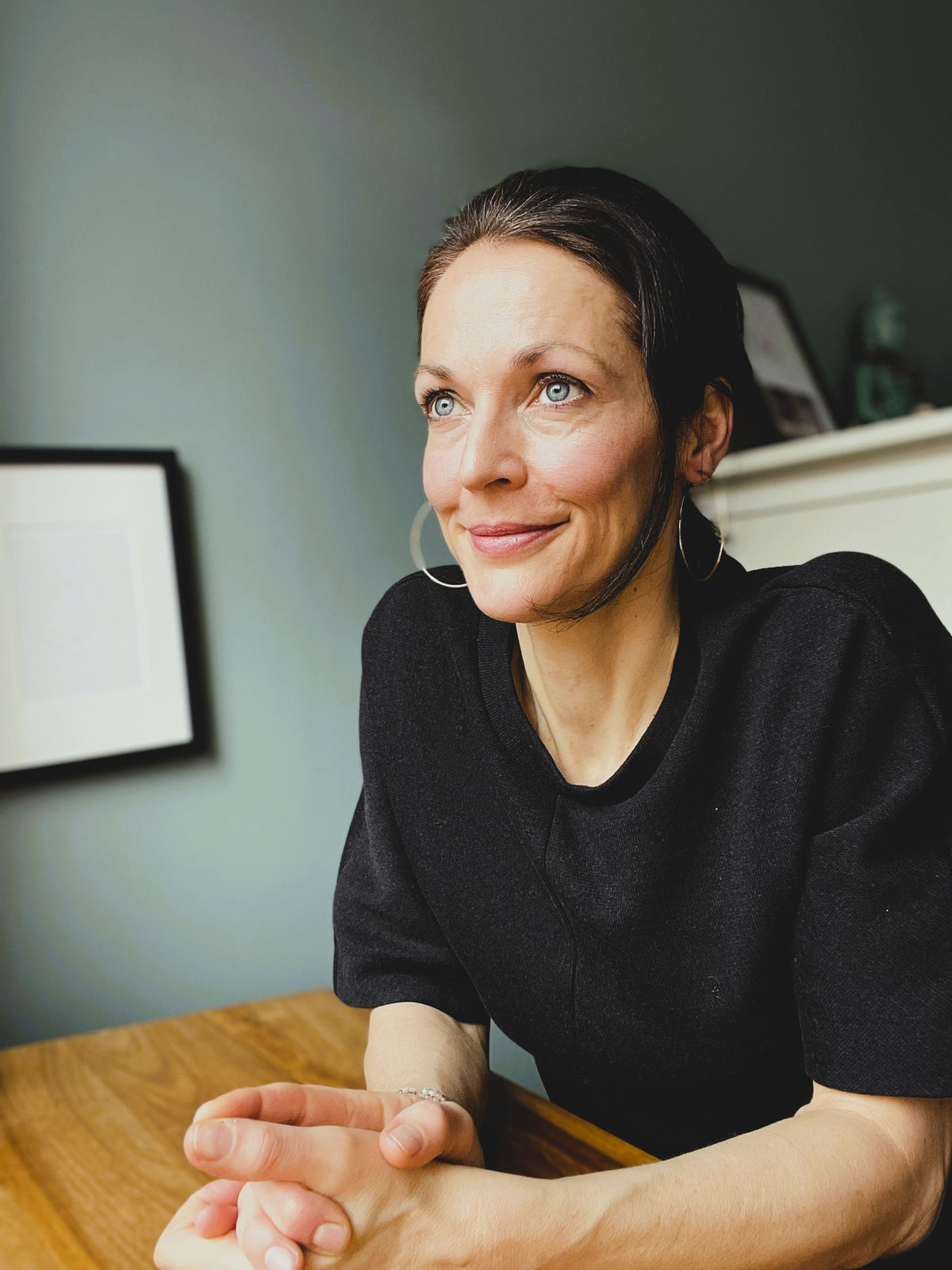 Regine Hüttebräuker sitzt mit gefalteten Händen am Holztisch vor einer salbeigrünen Wand und schaut lächelnd zur Seite.