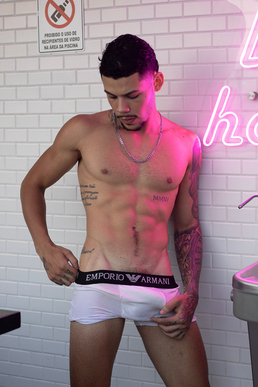 Homem tatuado sem camisa ajusta a cueca em banheiro branco com néon rosa