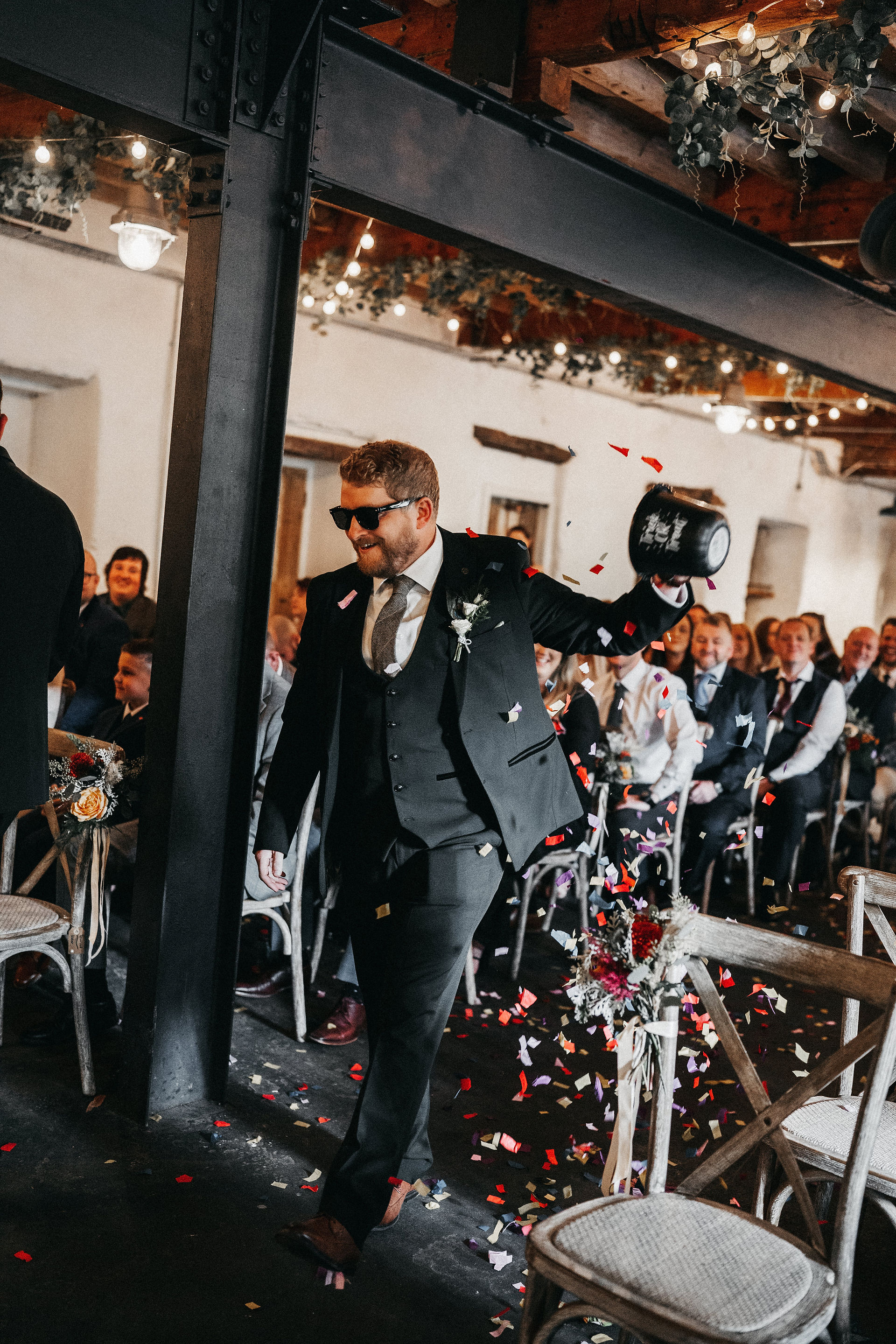 groom and groomsmen walking down the aisle throwing confetti and dishing out sweets and drinks to guests who are cheering and clapping
