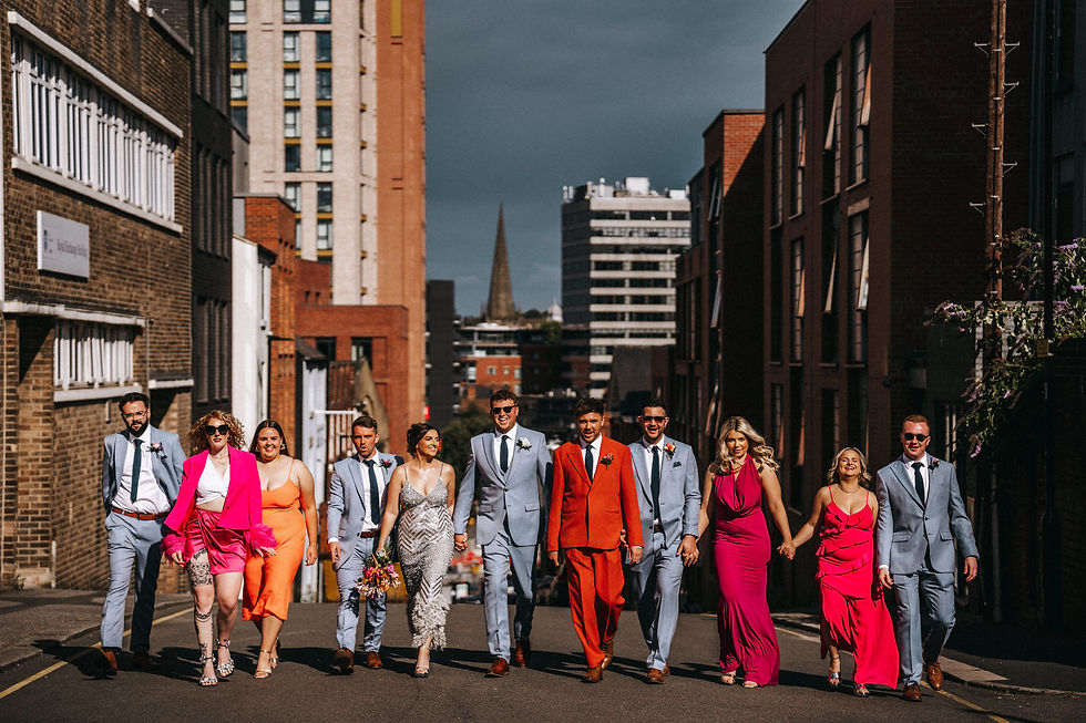 bride in silver dress and groom in light blue suit in a line with bridesmaids in oranges and pinks and groomsmen in matching suits all walking towards the camera with sheffield cityscape in the background