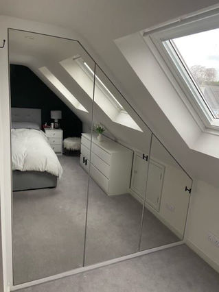 Loft eave fitted wardrobe extending from floor to ceiling with custom-built drawers, hanging rails and shelving, finished with glass doors by New Heights Carpentry in Reigate, Surrey. Designed for style and functionality.