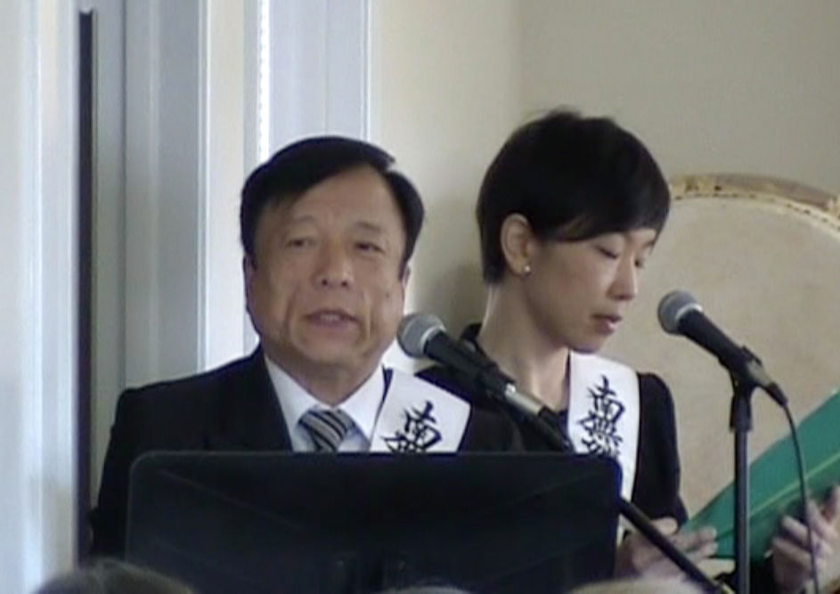 Two people in formal attire speak into microphones. They wear sashes with black text. One holds a green folder. The setting is indoors.