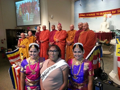 People in traditional attire and sashes stand in a hall with a Buddhist statue. Monks are in the background with colorful flags and banners.