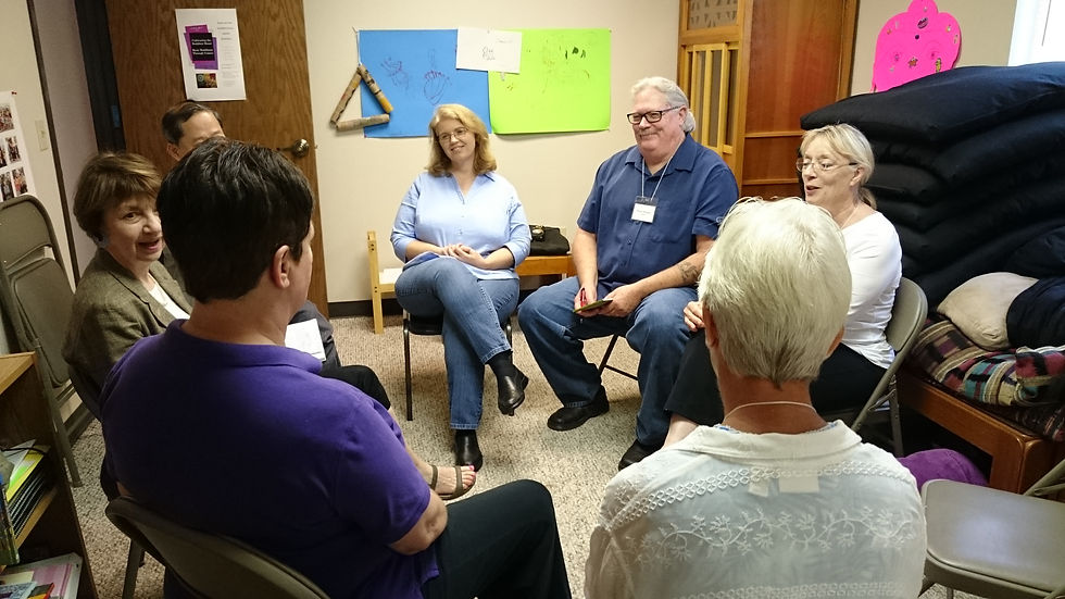 A group of seven people sit in a circle in a cozy room with colorful posters in the background, engaged in a friendly discussion.