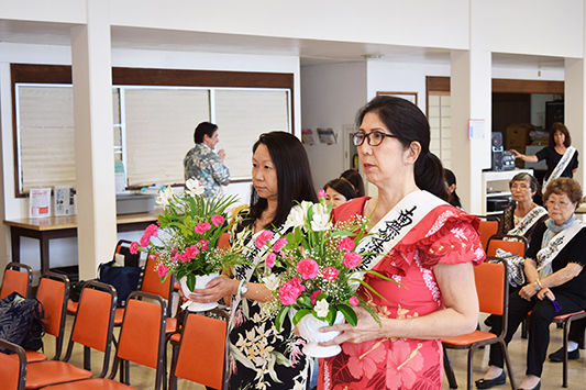 Two women in floral sashes hold bouquets of pink and white flowers in a room with orange chairs. Others sit in background, looking attentive.