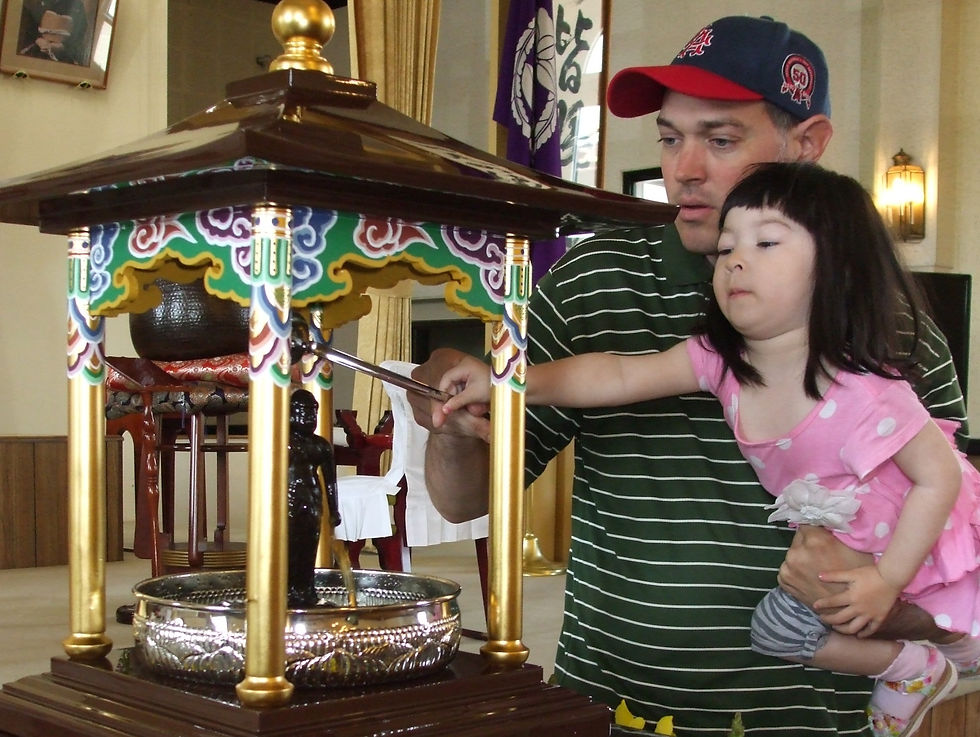 Man holds girl in pink as she pours water over a small statue under a colorful canopy in a temple. Interior with warm lighting.