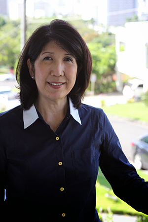 Woman in navy shirt smiling outdoors against a blurred green and urban background, conveying a calm and welcoming mood.