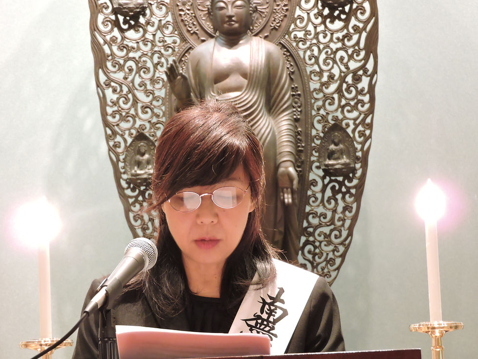 A person reads at a podium with a microphone, flanked by candles, in front of a Buddha statue. They wear glasses and a sash with Japanese text.