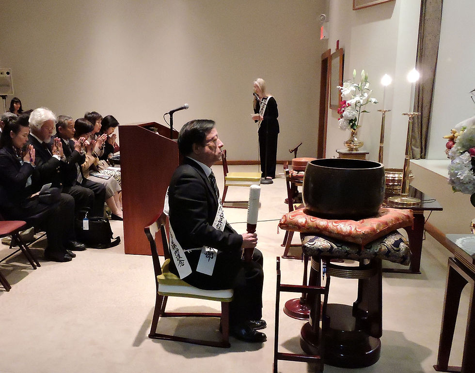 Man in suit kneeling by a large bowl; people seated, hands in prayer. Woman at podium, floral arrangements; solemn ceremony setting.