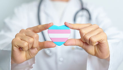Doctor wearing a white coat, holding up a heart-shaped badge which displays the trans flag.