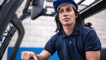 A muscular non-binary person of colour driving a forklift indoors. They are wearing a blue hard hat and a blue polo shirt. They have long black hair.