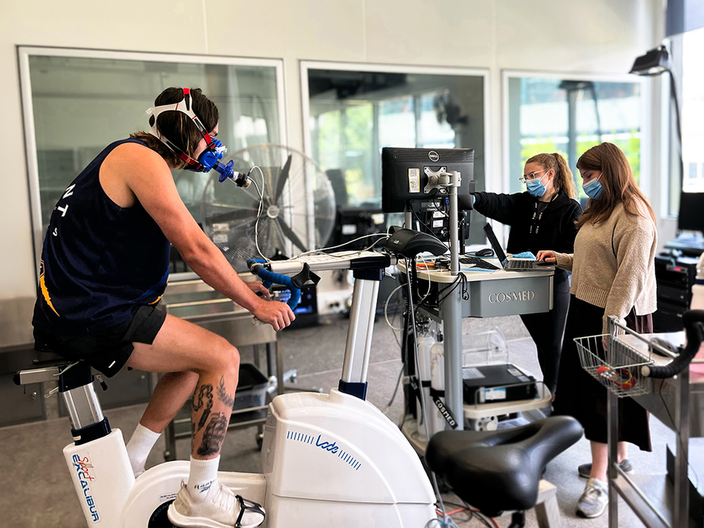 A person cycling on a stationary bike while two doctors stand nearby, monitoring the person's cycling