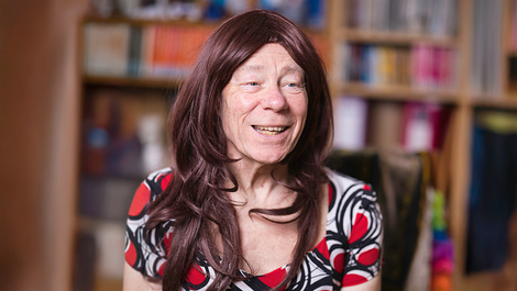 An elderly, light-skinned non-binary person sitting indoors, smiling happily and looking into the distance. They are wearing a maroon-coloured, long-haired wig and a patterned dress comprised of red circles and black lines against a white background. Bookshelves, books, and wooden walls are visible in the blurred background.