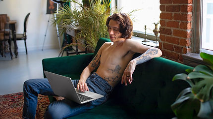A trans man with top surgery scars sitting on his couch, looking at his laptop