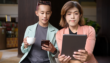 A non-binary person drinking coffee and looking at their phone, sitting next to a non-binary transgender woman who is looking at her digital tablet