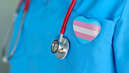 A close-up photo of a nurse's blue scrubs, focusing on a badge which is pinned to their front pocket. The badge has white, pink, and blue stripes, representing the trans flag. The nurse is also wearing a red stethoscope around their neck.