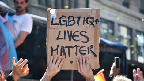Two hands holding up a cardboard sign during a pride parade, with other people and buildings visible in the blurred background. It is a sunny day. Text on the cardboard, written in black marker, reads, "LGBTIQ+ LIVES MATTER," followed by a love heart.
